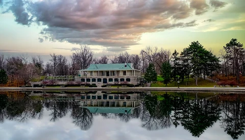 Classical pavilion with columned portico reflected in still water at dusk under dramatic clouds.