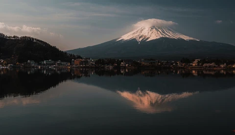 Snow-capped volcanic mountain reflected in still lake water at dawn, with town buildings along shoreline.
