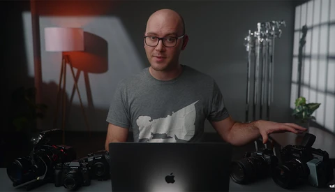 Male photographer at desk with laptop surrounded by professional cameras and studio lighting equipment.
