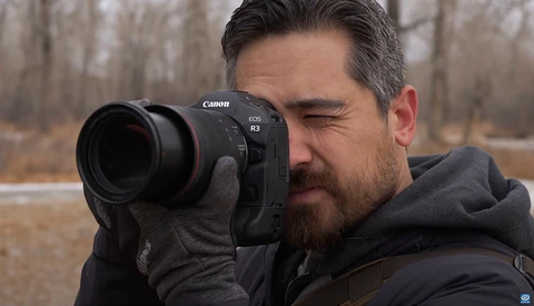 Photographer holding Canon EOS R3 camera with telephoto lens, eye pressed to viewfinder.