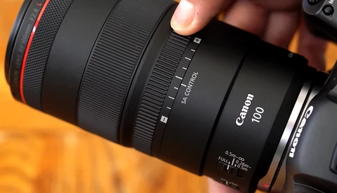 Canon RF 100mm macro lens held in hand against warm orange background.