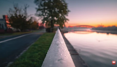 Concrete railing extending toward a canal at sunset with trees and buildings silhouetted against pink and orange sky.