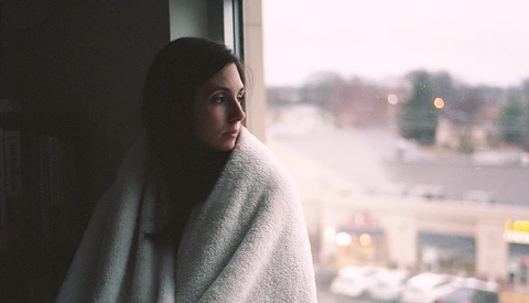 Woman wrapped in a knit blanket gazing out a high-rise window at an urban street below.