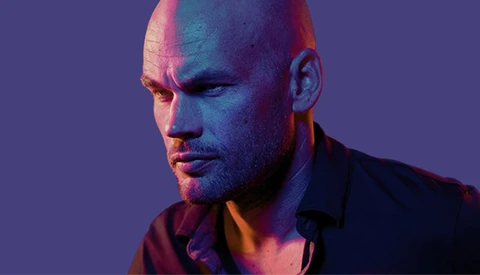 Bald man with facial hair against purple background with warm and cool lighting.