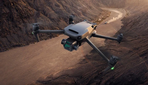 Drone hovering above an arid canyon landscape with a winding dirt road and vehicles below.