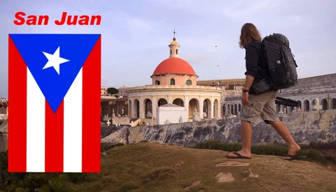 Traveler with backpack standing on rocky terrain overlooking a colonial dome building with Puerto Rican flag displayed on left.