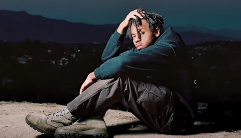 Young man sitting on ground at dusk with head resting on arm, wearing teal jacket and dark pants.