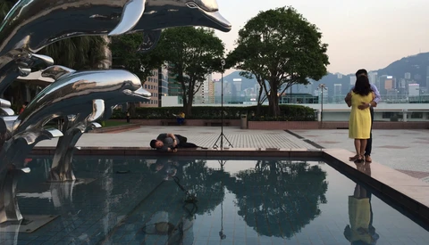 Photographer lying on wet pavement to shoot reflections in still water, with woman in yellow dress posing nearby.