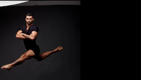 Male dancer in black outfit performing a horizontal leap against dark background.