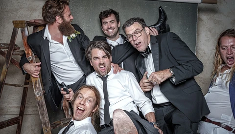Group of six men in formal attire celebrating exuberantly at an indoor event.