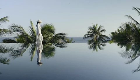 Woman in white linen dress standing on a mirror-like pool reflecting palm trees and sky.