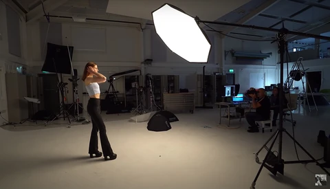 Female model posing during a studio photoshoot with professional lighting setup and crew members present.