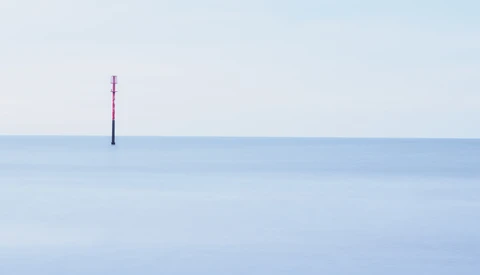 A single red and black pole stands isolated in calm, shallow blue water against a clear horizon.