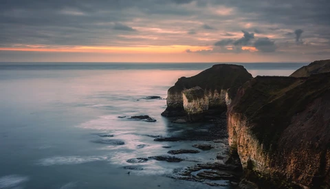 Coastal cliffs overlooking rocky shoreline at sunset with smooth water and dramatic sky.