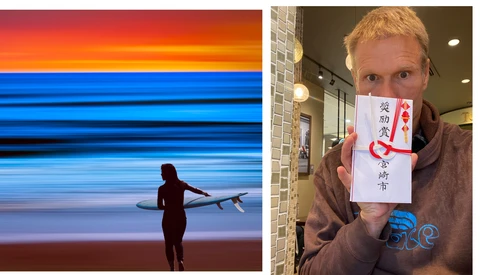 Surfer in wetsuit carrying board at sunset beach with vibrant orange and blue sky.