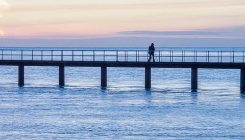 Solitary figure standing on a long pier extending into calm blue water at twilight.