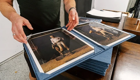 Hands holding open a blue cloth portfolio displaying two vintage black and gold photographs.