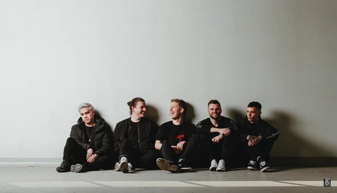Five men in dark clothing sitting against a white wall with dramatic shadows.