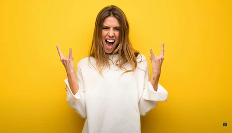 Woman in white shirt making rock gesture with both hands against bright yellow background.