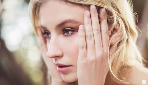 Blonde woman in profile with hand near face, soft natural lighting and blurred background.
