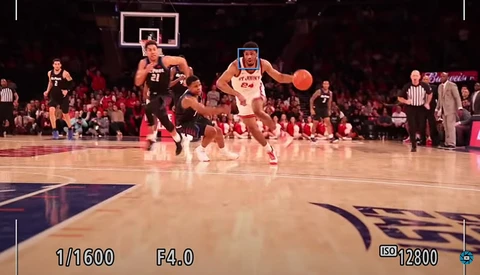 Basketball player in white jersey dribbling ball during live game with packed arena in background.