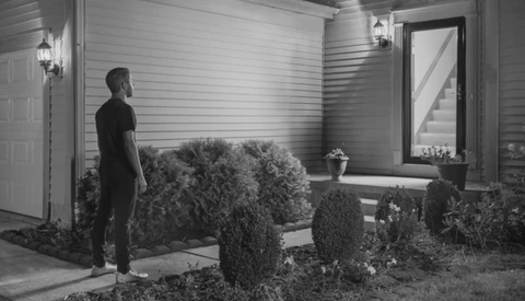 Man in dark clothing standing on a residential porch at night, surrounded by landscaped shrubs.