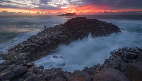 Rocky coastal headland with a solitary figure silhouetted against a dramatic sunset sky over the ocean.