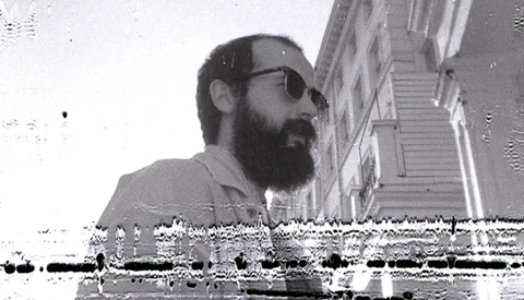 Bearded man wearing sunglasses and light-colored shirt, photographed in profile against urban staircase backdrop.