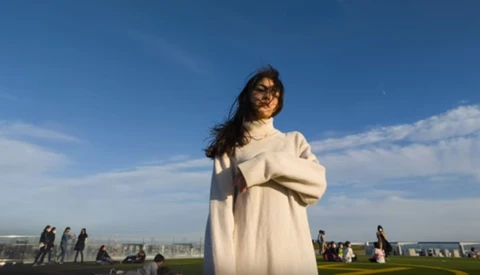 Woman in cream sweater standing outdoors against blue sky with distant cityscape.