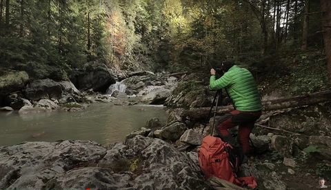 Photographer in bright green jacket operating a tripod-mounted camera beside a flowing forest stream.