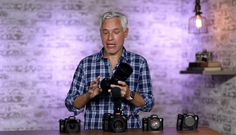 Man in blue plaid shirt holding camera equipment while standing behind four cameras on a table.