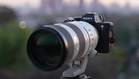 Sony α7 mirrorless camera with white telephoto lens mounted on tripod against blurred urban background.