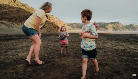 An adult and three children playing together on a dark sand beach backed by tall cliffs.