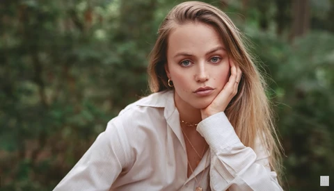 Woman with long blonde hair wearing white shirt, resting chin on hand against blurred green foliage background.
