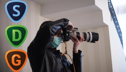 Photographer wearing a mask and black jacket holding a DSLR camera with telephoto lens to their eye.