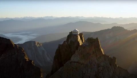 Mountain monastery perched on a rocky peak at sunrise, with layered mountain ranges and morning mist in the valley below.