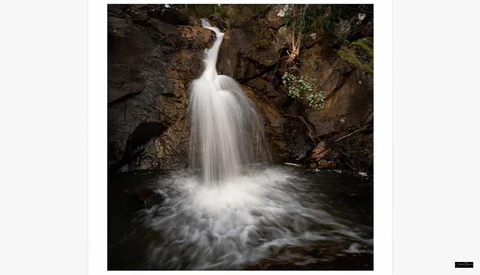 Small waterfall cascading into a rocky pool surrounded by dark stone walls and moss-covered vegetation.