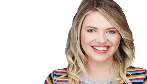 Portrait of a smiling woman with wavy blonde hair wearing a colorful striped top against a white background.