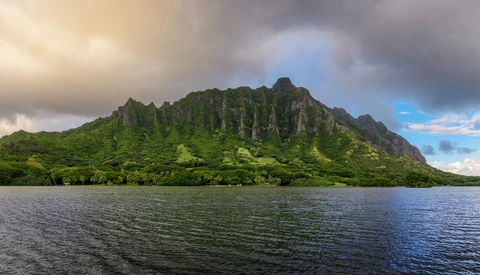 Lush green mountain ridge rising from calm blue water under partly cloudy sky.