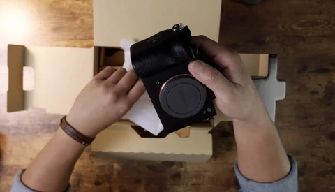 Hands holding a mirrorless camera body with lens cap removed, photographed from above on a wooden surface.