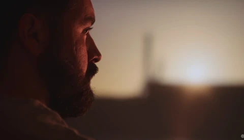 Bearded man in profile silhouette against warm sunset or golden hour light with blurred cityscape behind.