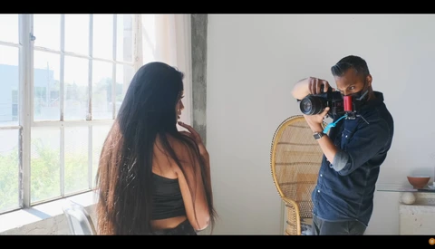 Photographer holding a professional video camera while filming a woman with long dark hair in a bright studio with large windows.