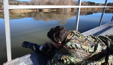 Photographer shooting with a telephoto lens from a wooden pier overlooking a calm lake with bare trees reflected in the water.