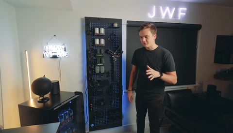Man in black t-shirt gesturing while speaking next to server equipment and lighting setup in studio.