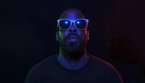 Man wearing illuminated blue-framed glasses against dark background with colored gel lighting.
