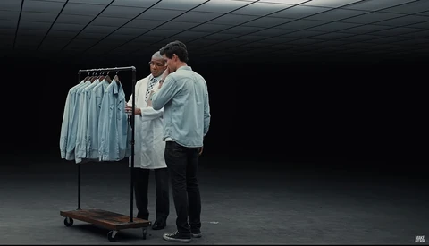 Two people examining garments on a rolling rack in a minimalist studio space with dark walls.