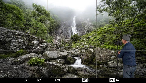 Photographer standing on mossy rocks beside a flowing stream with a misty waterfall visible in the forested background.