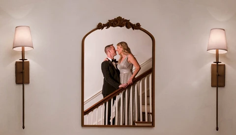 Couple in formal attire kissing on a staircase, reflected in an ornate arched mirror flanked by wall sconces.