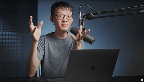 Young person wearing glasses speaking animatedly at a laptop with a studio microphone overhead.