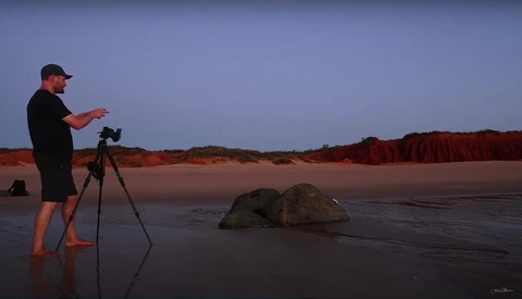 Photographer on a beach at dusk operating a camera on tripod, facing rocky outcrops in shallow water.